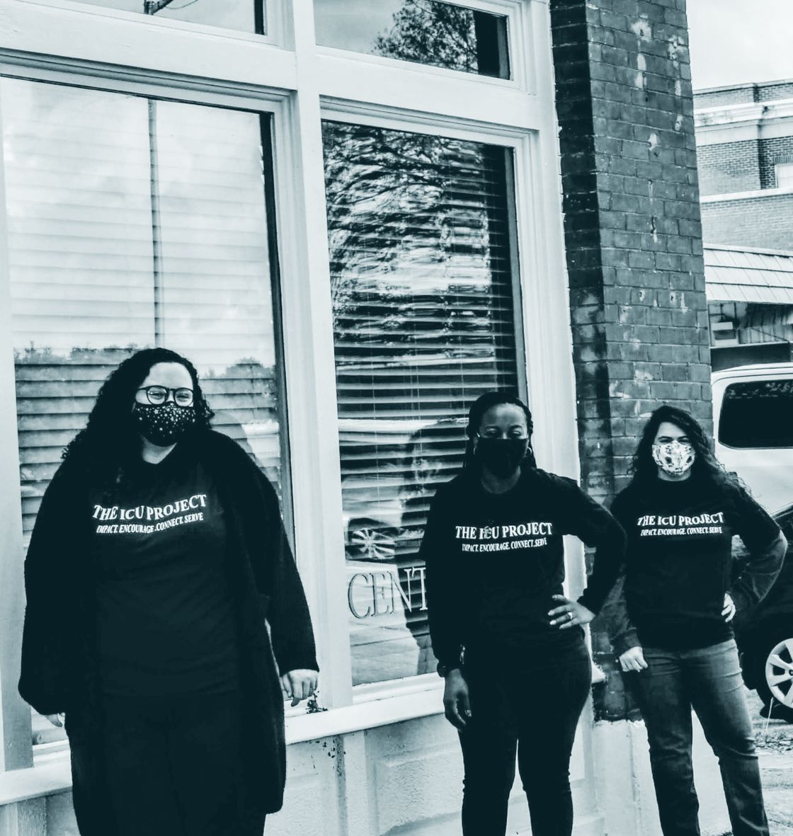 Three ICU Project members in masks and black shirts printed with The ICU Project and Impact, Encourage, Connect, Serve, standing outside a storefront.